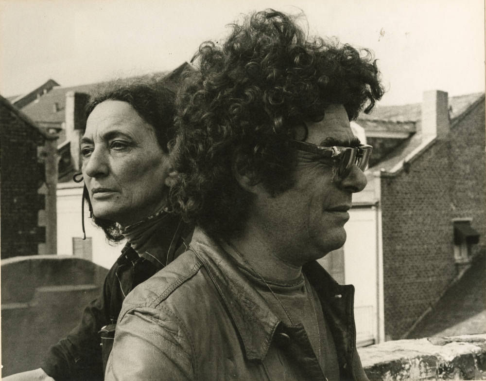 Two people stand outside, one wearing glasses and a leather jacket, the other with long hair and a scarf. They look in opposite directions, with buildings visible in the background. The photo is in black and white.