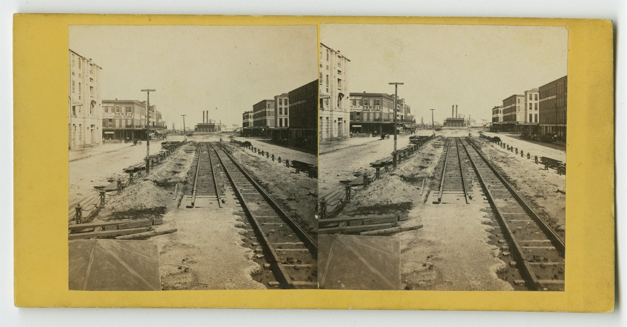 A vintage stereoscopic image showing a city street with railroad tracks in the center, flanked by buildings on both sides. The road is unpaved and there are stacks of materials alongside the tracks.