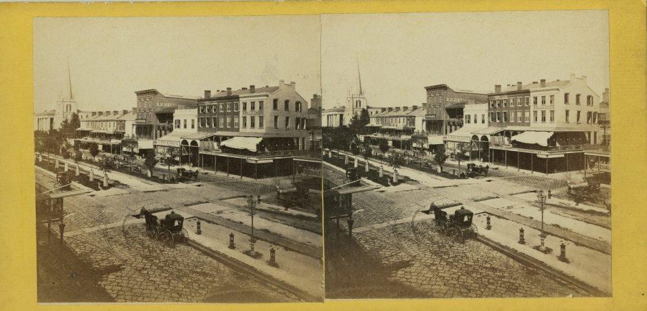 A sepia-toned stereograph of a bustling 19th-century street scene. Horse-drawn carriages travel along a cobblestone road lined with brick buildings and storefronts. A church with a tall steeple is visible in the background.