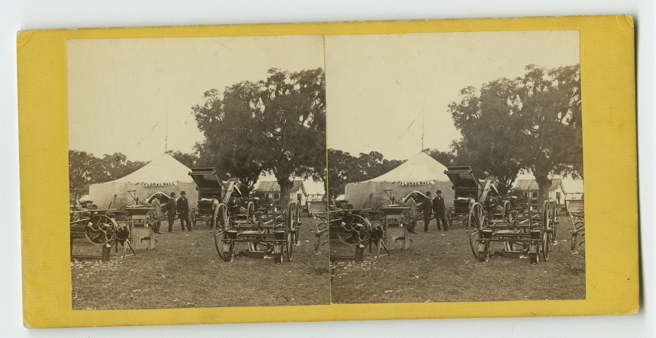 A vintage stereoscopic photograph depicts a field scene with several people standing near a tent labeled U.S. Sanitary Commission. There are cannons and equipment in the foreground, under a few sprawling trees.