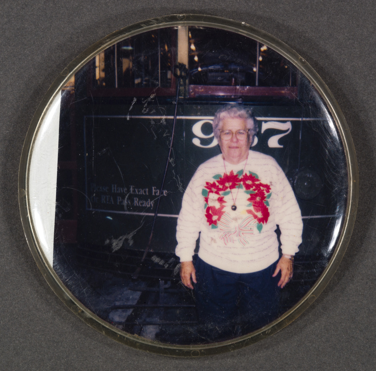 An elderly woman with short white hair stands smiling in front of a green vintage streetcar numbered 967. She is wearing a white sweater with a red floral pattern on the front.