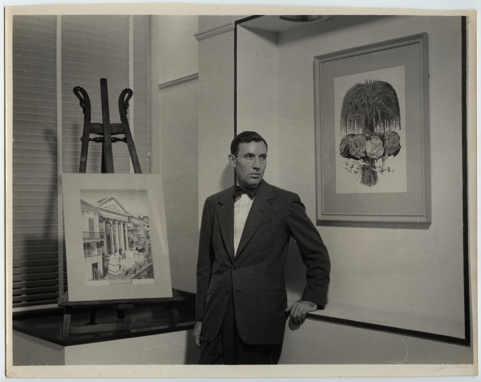 A black and white phot of a man (Boyd Cruise) in a dark suit and bow tie standing in a galelry space. He is flanked on his right by an easle showing an illustration of a building, and a framed illustration of a floral arrangement on his left.