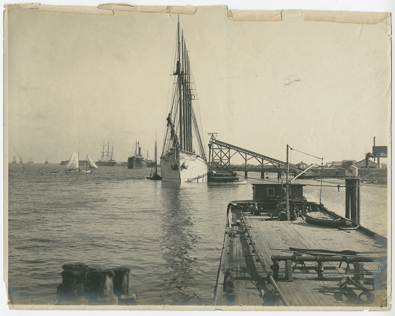 View of an industrial site or port, that show a variety of boats and ships on the water and a loading mechanism extending out over the water where a ship is moored.