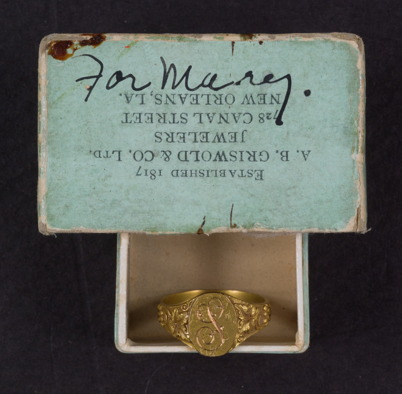 An ornate gold ring sits in an open, vintage jewelry box with a light blue interior. The inscription on the box reads For Mary, and its from a jeweler located in New Orleans, Louisiana. The top of the box is partially visible.