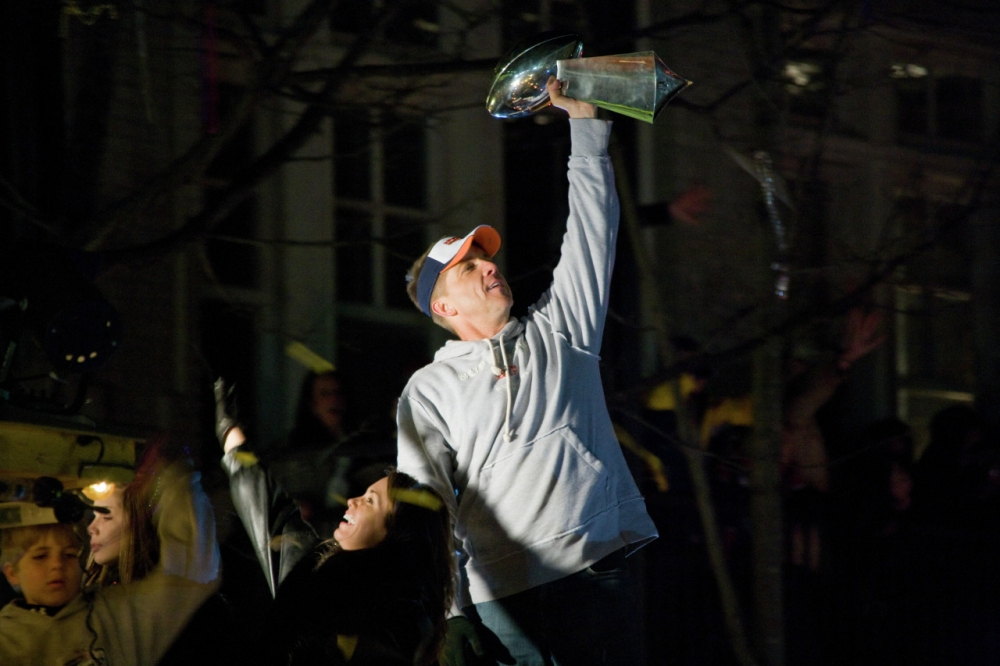 A person in a gray hoodie and baseball cap lifts a large trophy triumphantly. They stand among a cheering crowd, some with hands raised, under night lighting.