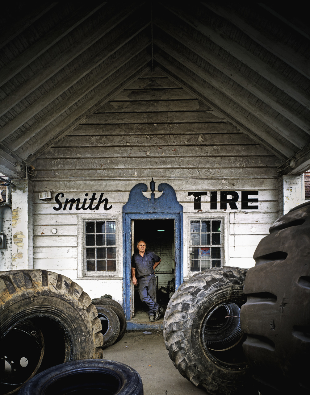 A man stands in the doorway of an old wooden building with Smith Tire written on the front. Large tires are scattered around the entrance, and the building has a steep, slanted roof and a blue-framed door.