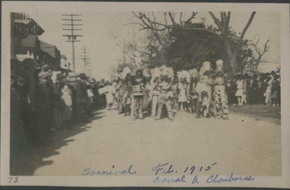 A black and white photo of a carnival parade from February 1915. People dressed in costumes walk down a street, lined with spectators on both sides. Handwritten text reads Carnival Feb. 1915 Canal & Claiborne.
