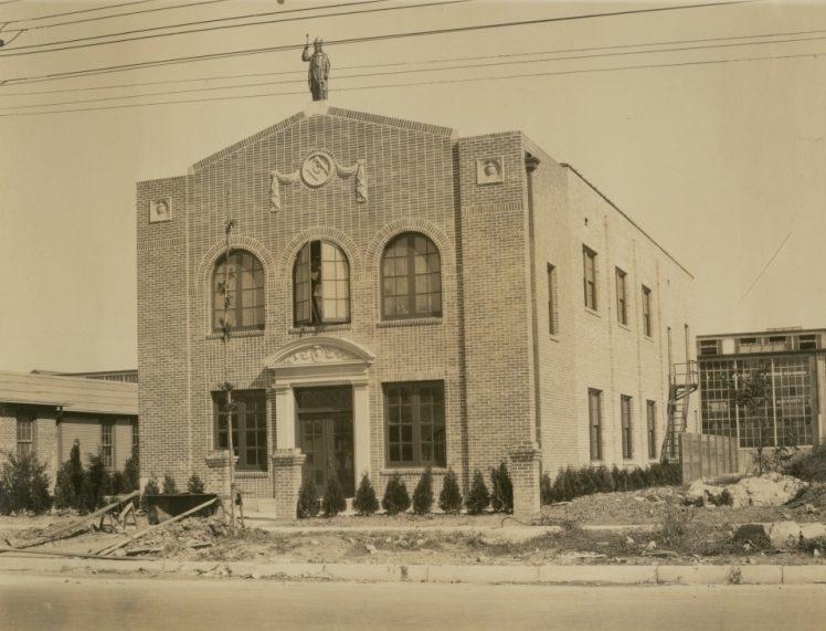 A vintage photograph of a two-story brick building with arched windows, a decorative figure on the roof, and a small garden in front. A street and power lines are visible in the foreground.