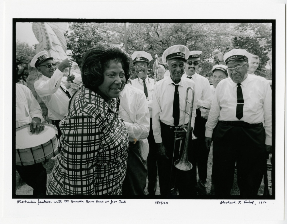 A group of jazz musicians pose outdoors, wearing white shirts and caps. Marian Anderson stands in the foreground, smiling in a patterned dress. A trombonist holds his instrument while others hold drums and a trumpet. Trees are visible in the background.