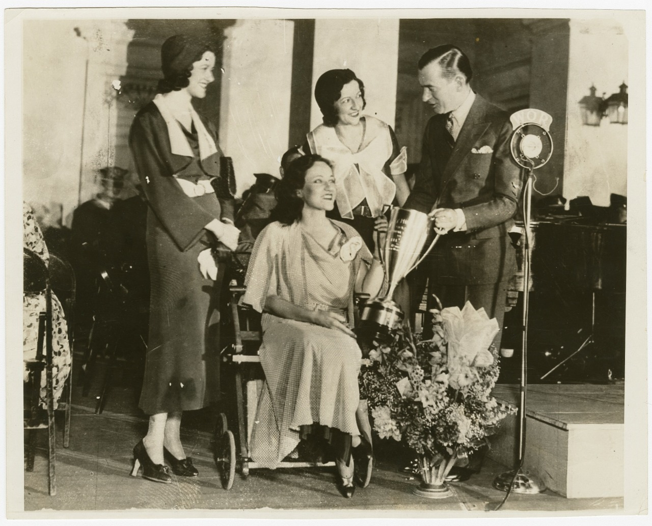 A black-and-white photo of four people, two women standing on the left, a woman seated in the center, and a man standing on the right near a microphone. The man is handing a trophy to the seated woman. A large bouquet is in front of them.
