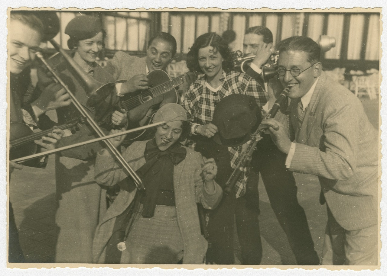 A vintage black and white photo of a lively group of men and women playing violins and a horn. Theyre smiling and wearing 1930s-style clothing, capturing a joyful musical moment outdoors.
