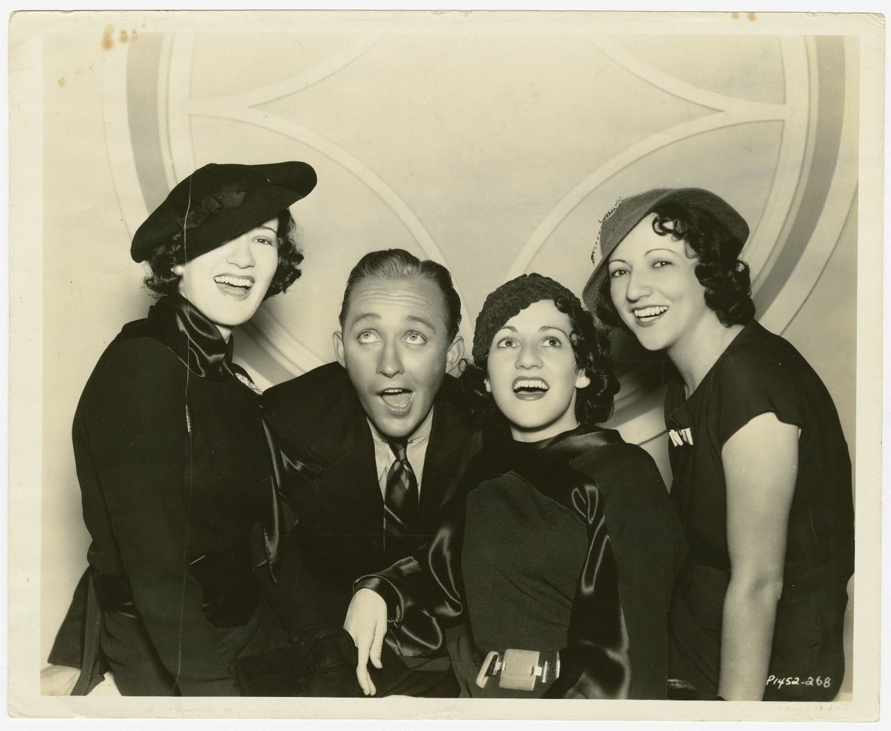 Black and white photo of a man and three women smiling and laughing, all dressed in fashionable 1930s attire, with hats and dark clothing, against an art deco style backdrop.