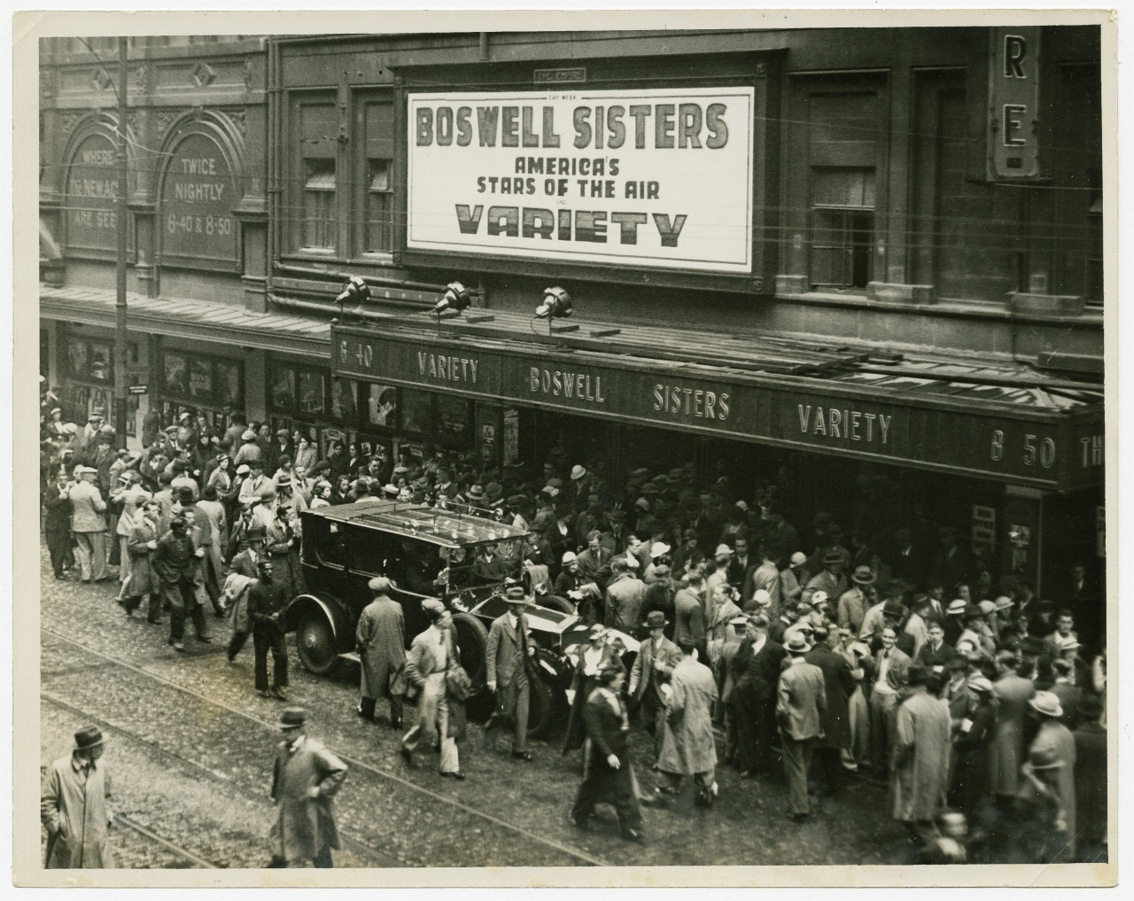 A busy crowd gathers outside a theater showing the Boswell Sisters Variety act. The marquee reads Americas Stars of the Air with people lining up and walking around the street, some carrying umbrellas.