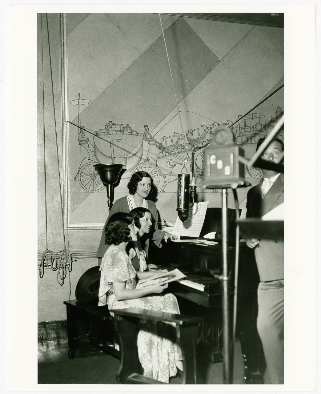 Three women in 1930s attire are gathered around a piano, with one playing and the others singing. A man operates a microphone and camera, recording the scene. The background features a mural of a horse-drawn carriage.