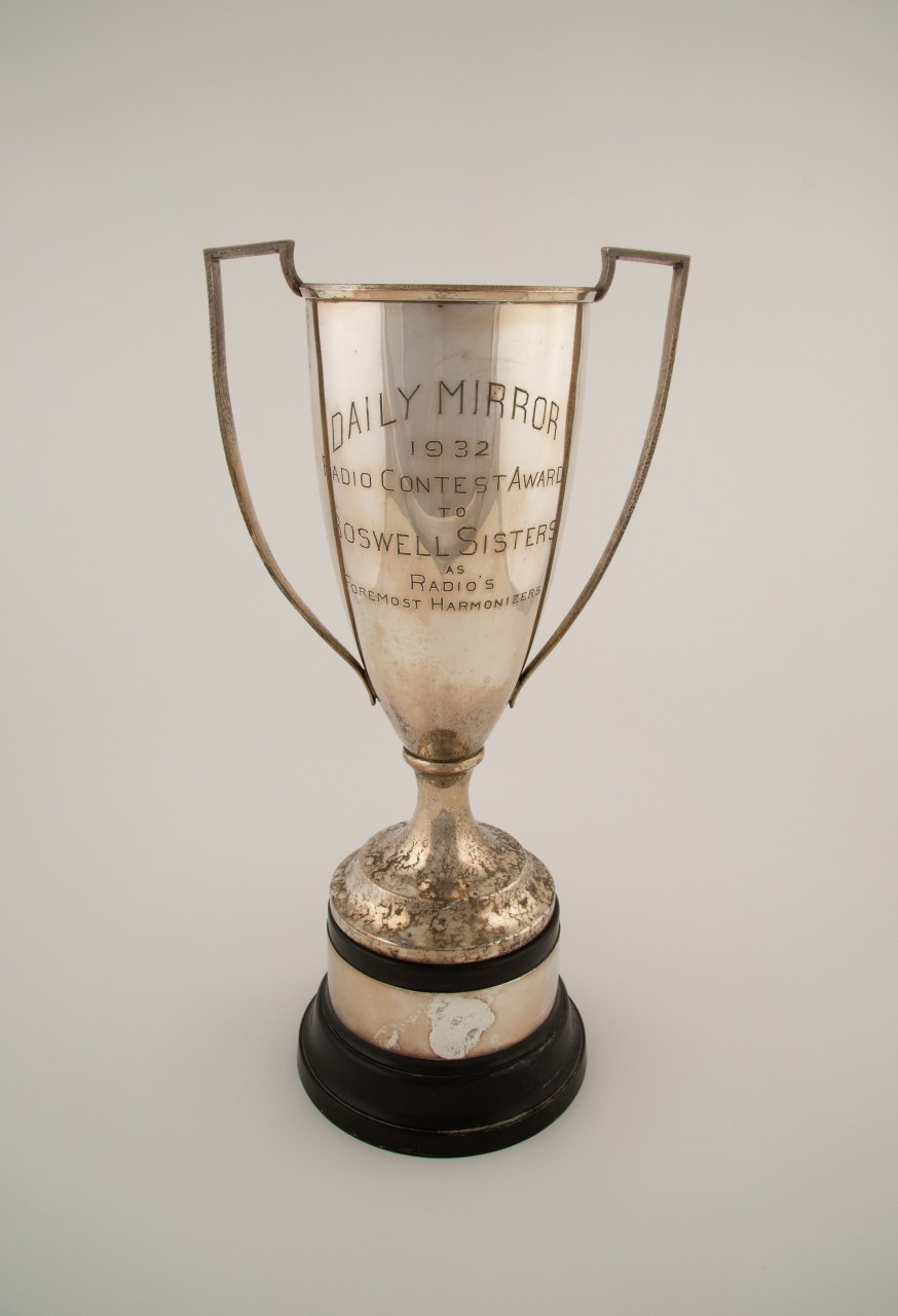 A vintage silver trophy with two handles, engraved with Daily Mirror 1932 Radio Contest Award to Boswell Sisters Radios Foremost Harmony Crew. It stands on a black base.