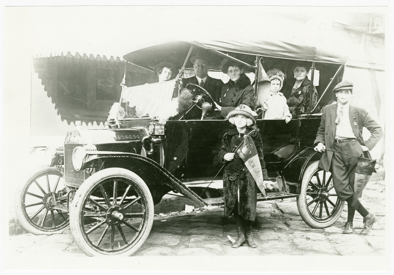 A vintage black-and-white photo showing a family seated in an open-top early 20th-century car. Two children stand in front, wearing period clothing. The scene captures a moment from a bygone era.