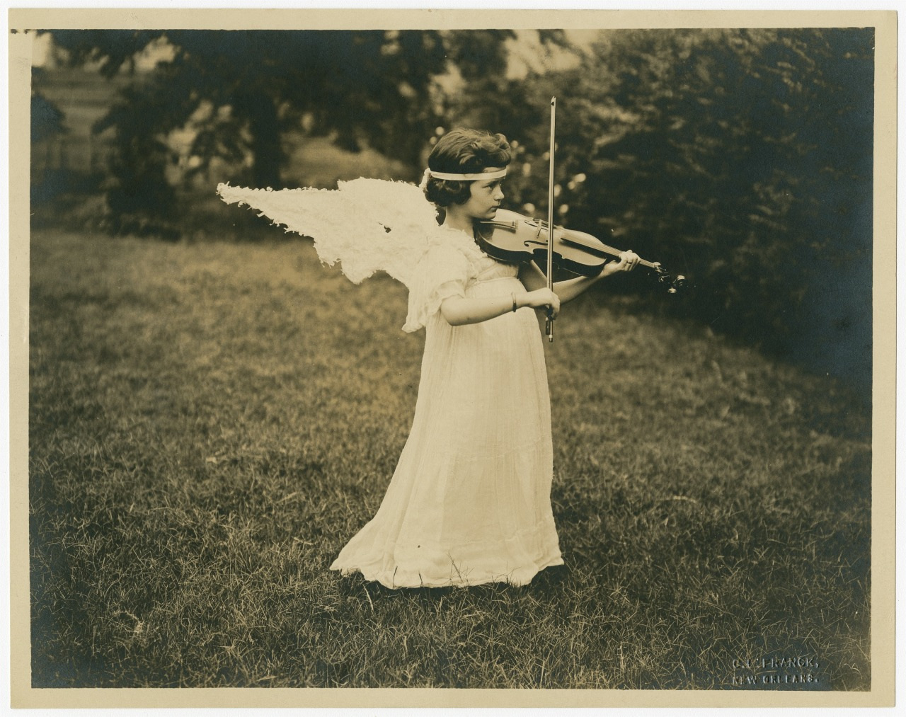 A young girl dressed as an angel plays the violin in a grassy outdoor setting. She wears a long white dress and angel wings, with a headband in her hair, creating a serene and enchanting scene.