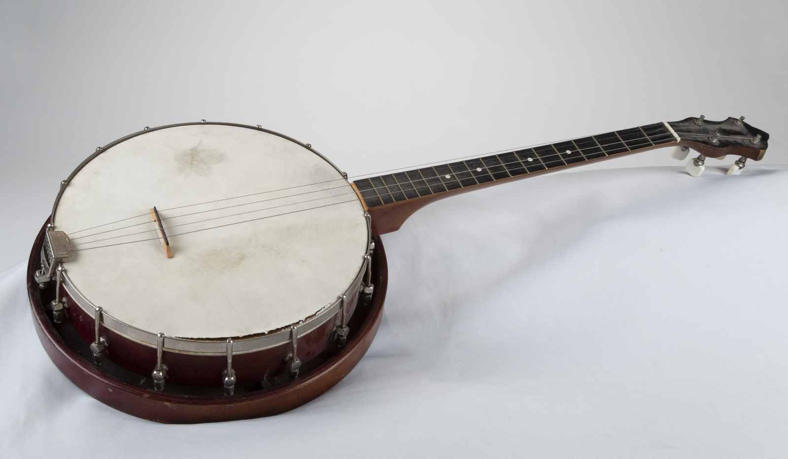 A vintage banjo with a wooden body, white drumhead, and four strings, resting on a white surface. The instruments metal brackets and tuning pegs are visible, giving it a classic and rustic appearance.