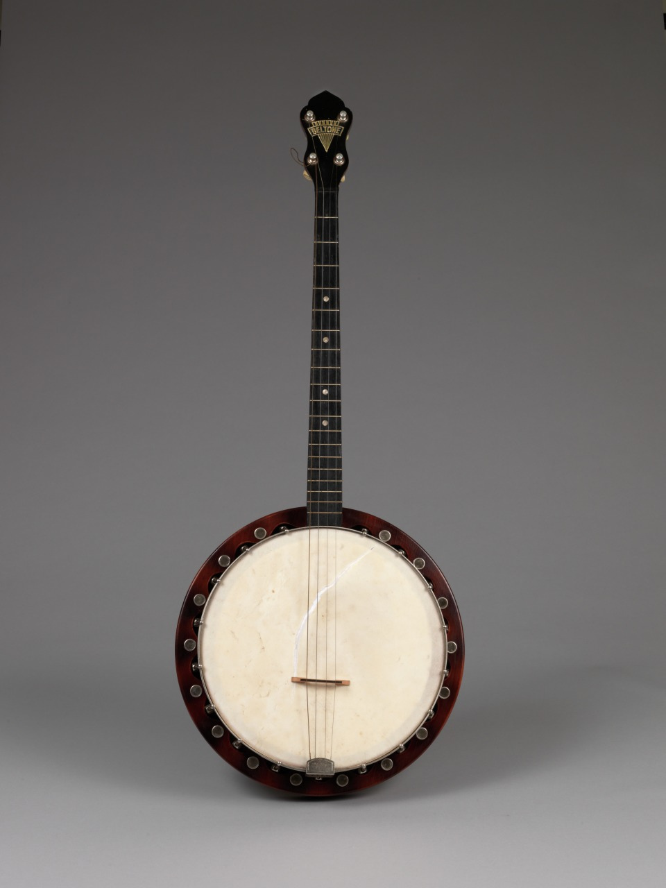 A four-string banjo with a dark neck and headstock, featuring metal tuners and a round body with a white drumhead. The body is bordered with a series of brackets connecting the drumhead to the frame. The background is a plain gray color.
