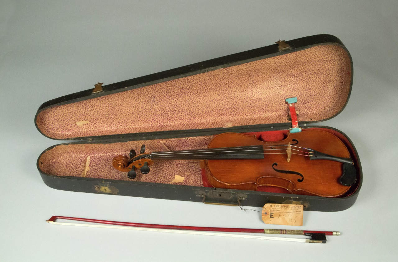 An open violin case with a violin inside, showcasing a worn brown body and scroll. The case is lined with pink fabric. A bow is placed beside the case, and a small tag is attached inside.