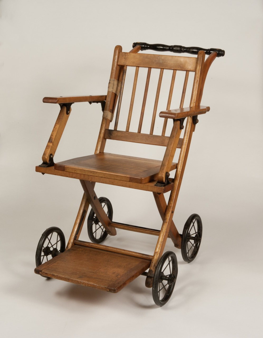 A vintage wooden wheelchair with black metal wheels and a slatted backrest is shown against a plain background. The design includes armrests and a footrest, combining wood and metal elements.