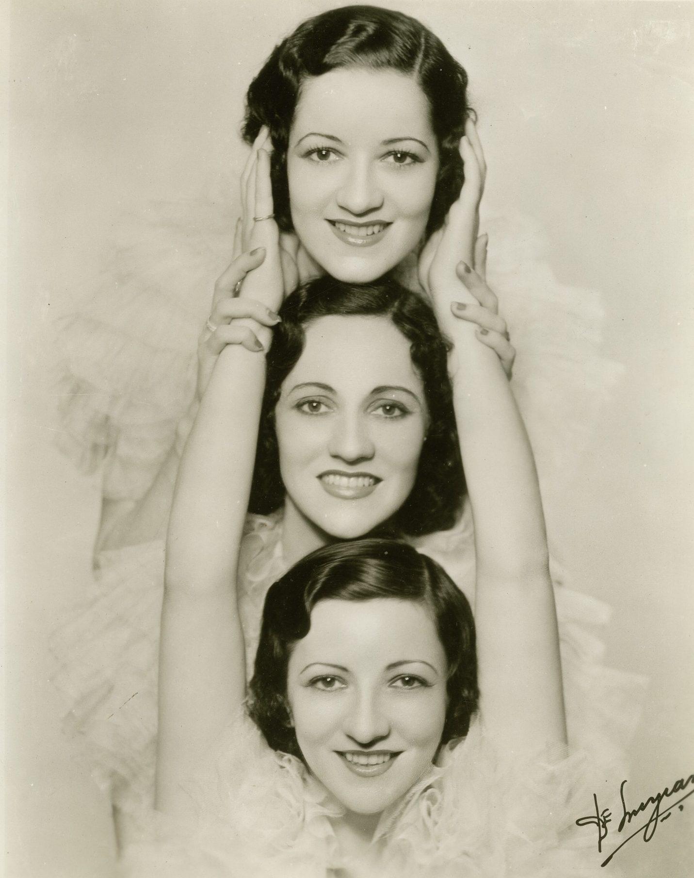 Three women are posed in a vertical line, smiling. Each woman rests her hands on the shoulders of the one above her. They have short, wavy hair and are wearing ruffled outfits, set against a plain background.