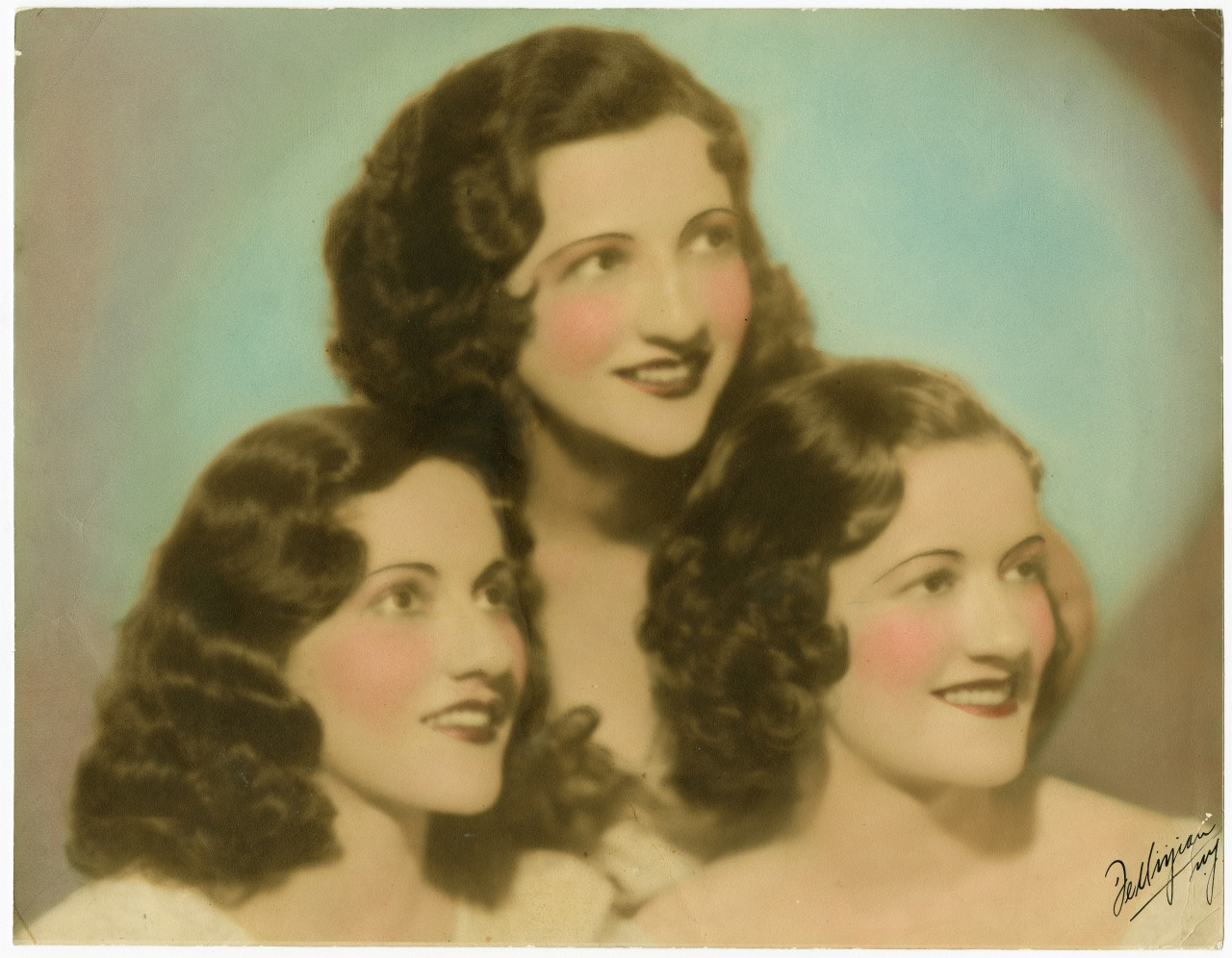 A vintage portrait of three women with wavy dark hair and bright smiles. They are wearing light-colored clothing and have rosy cheeks, against a softly blurred light background.