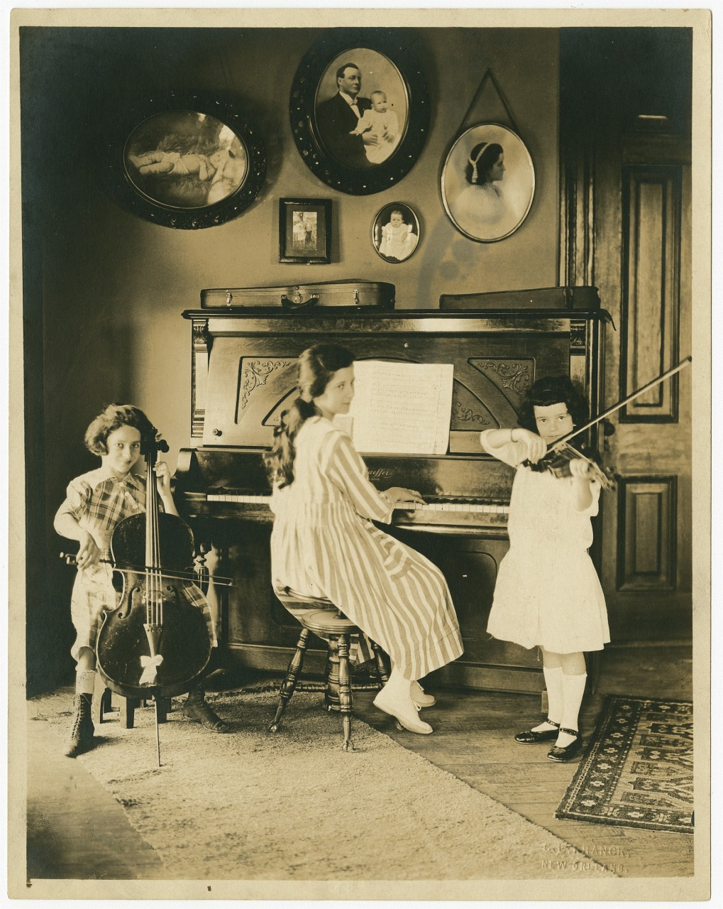 Three children play music in a room with a vintage piano and string instruments. One plays the piano, another the violin, and a third the cello. Framed portraits hang on the wall above, and they are dressed in early 20th-century attire.