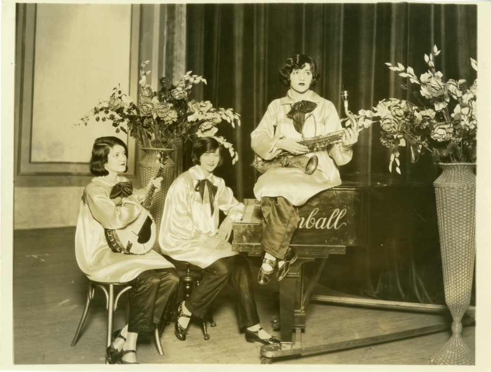 Three women sit and stand around a piano in a vintage setting. One holds a banjo, another a saxophone, and the third leans on the piano. They wear matching satin outfits with bows. Large flower arrangements frame the background.