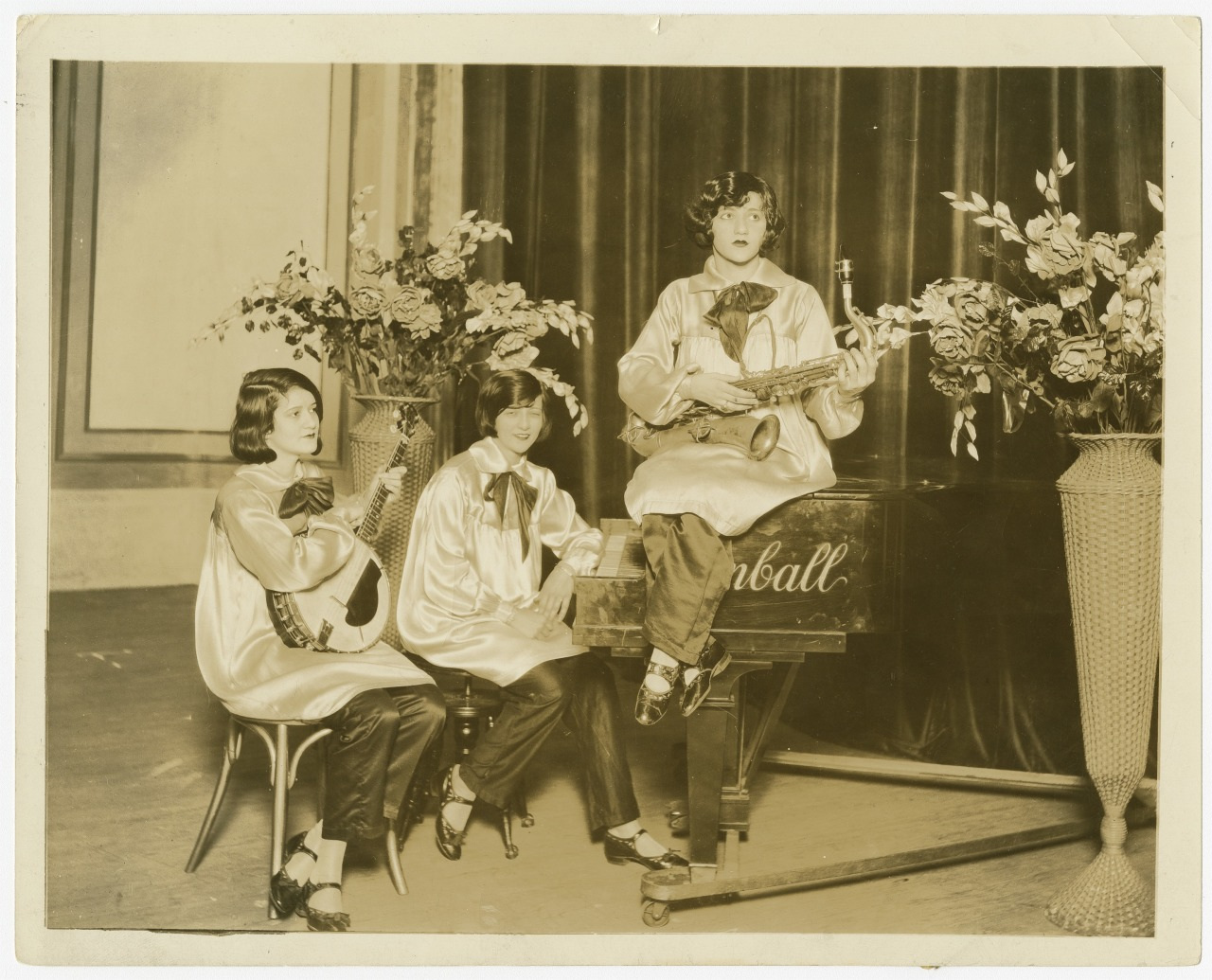 Three women in matching satin outfits pose with musical instruments in front of a piano. One holds a saxophone, another has a banjo, and the third is seated with a piano. Floral arrangements decorate the stage.