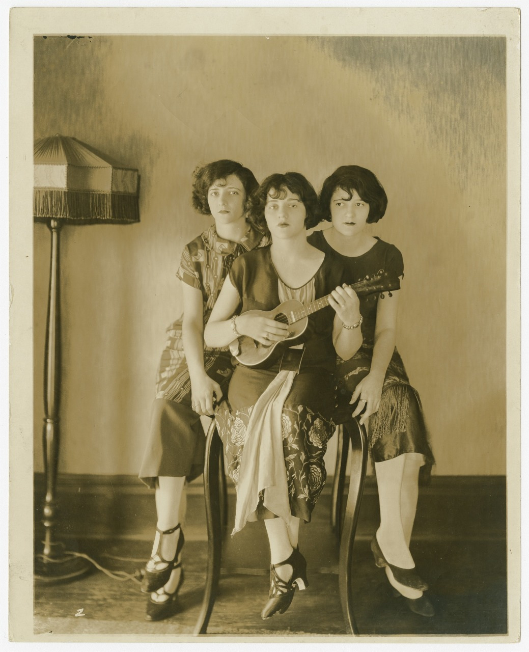 Three women sit and stand around a chair. The woman seated in the middle holds a small stringed instrument, likely a ukulele. They wear 1920s-style dresses and shoes. A fringed lamp stands to their left against a plain wall.