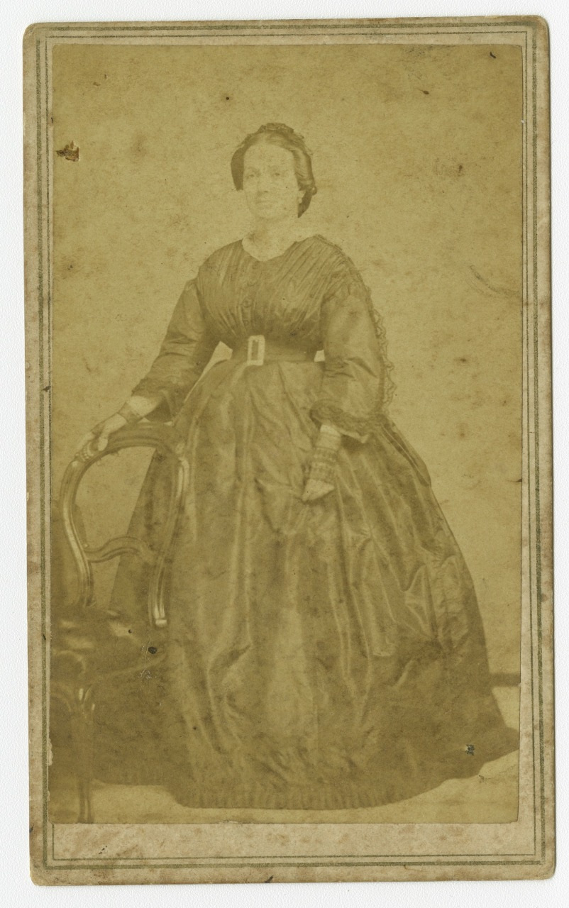 Sepia-toned vintage photograph of a woman in a long, dark, Victorian-era dress. She is standing with one hand resting on a wooden chair, and the image is slightly faded with age.
