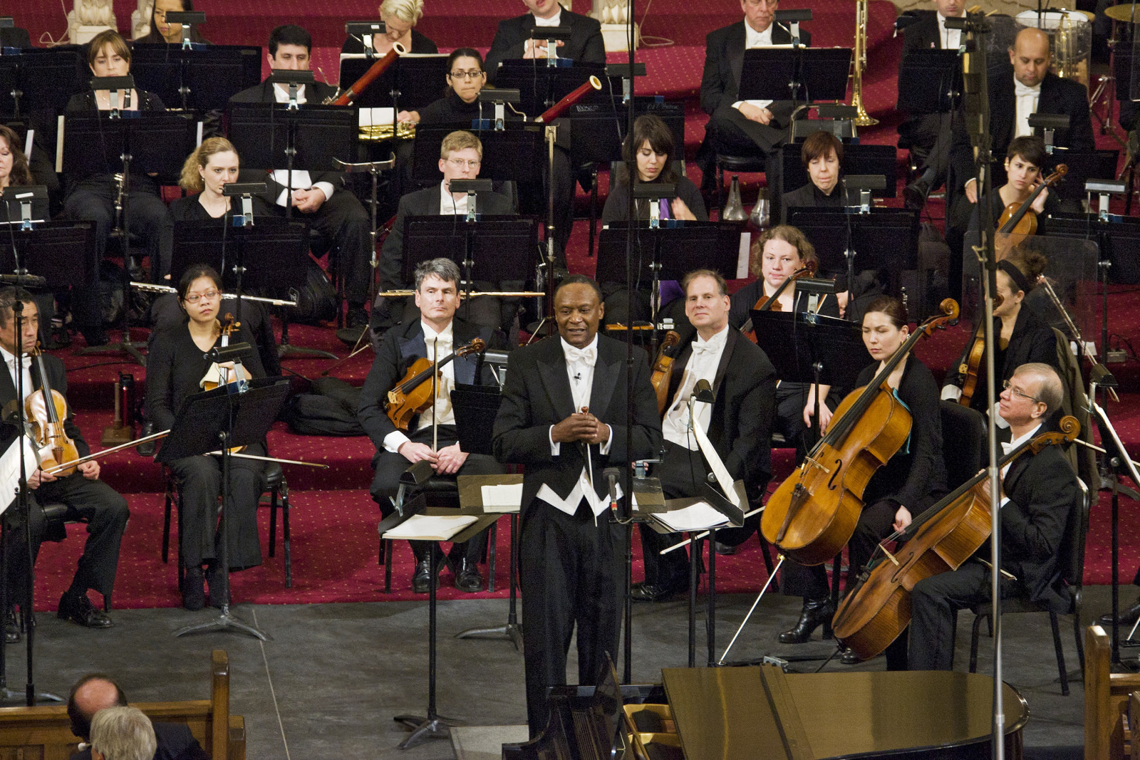 A conductor stands in front of a large orchestra, holding a microphone. The orchestra members are seated with their instruments, including violins, cellos, and wind instruments, against a maroon carpet background in a concert hall.
