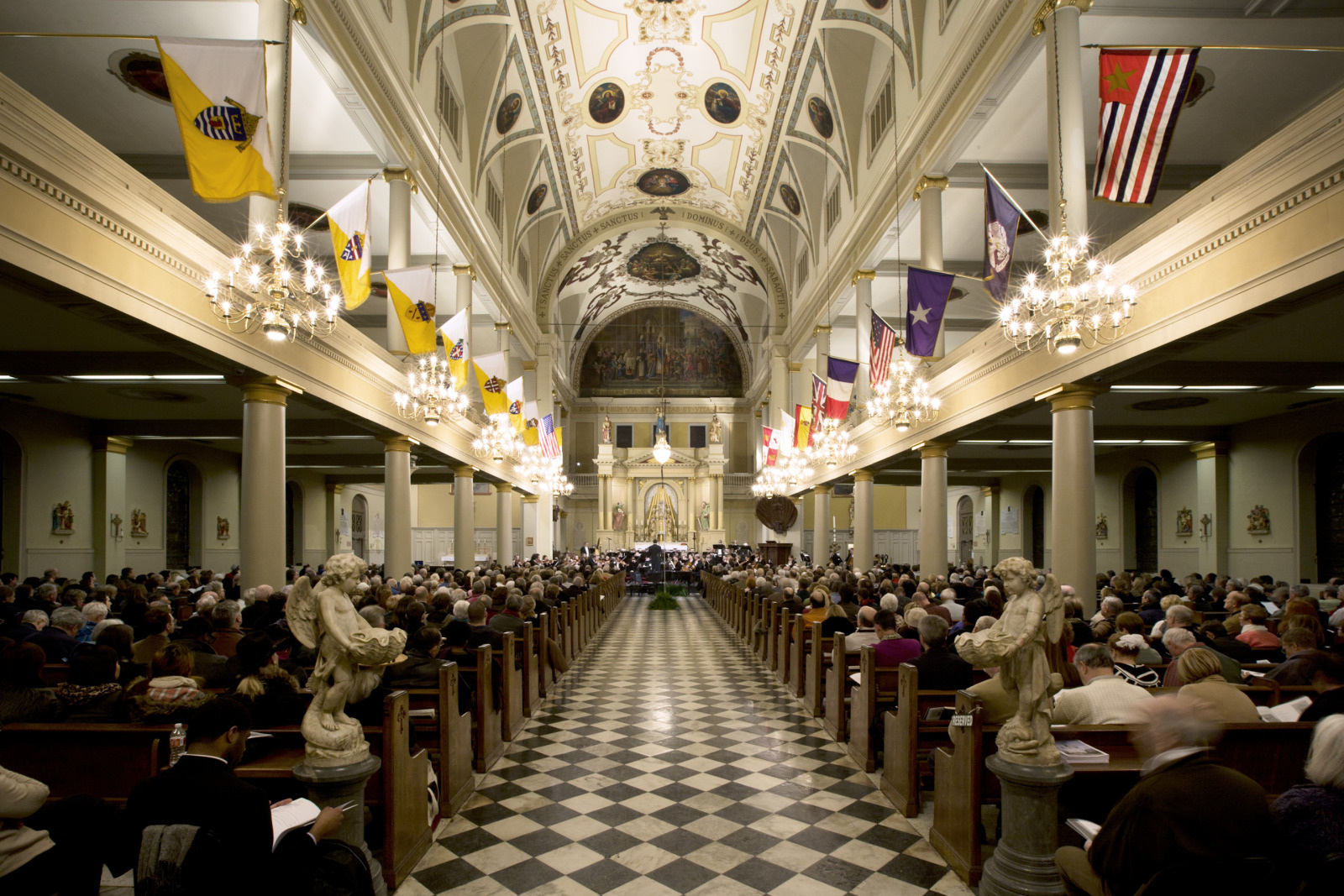 A large, ornate church interior with a checkered floor and chandeliers. The pews are filled with people attending a service, facing an altar adorned with flags and artwork. The ceiling features intricate designs and paintings.