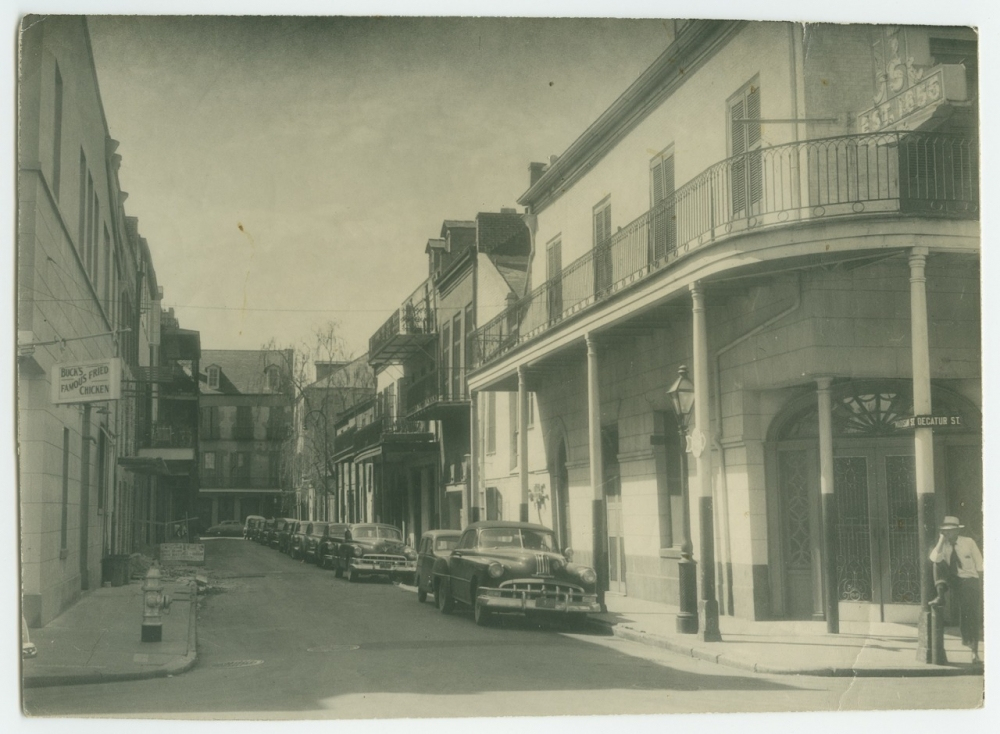 A vintage black and white photograph of a street lined with classic cars parked along the curb. The street is flanked by historic buildings with wrought iron balconies. A person is walking on the sidewalk, and signs are visible on the buildings.