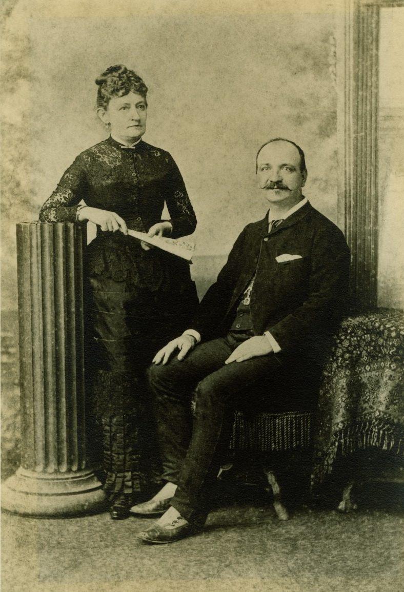 A vintage sepia-toned portrait of a woman standing and a man seated. The woman holds a fan and wears a dark dress, and the man has a mustache, wearing a suit. They pose in a room with a decorative column and textured curtain.