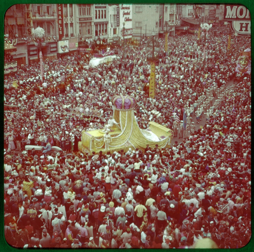 A vibrant aerial view of a large crowd gathered around a colorful parade float adorned with a giant purple and gold crown. The street is packed with people in festive attire, and the backdrop features historic storefronts and signage.