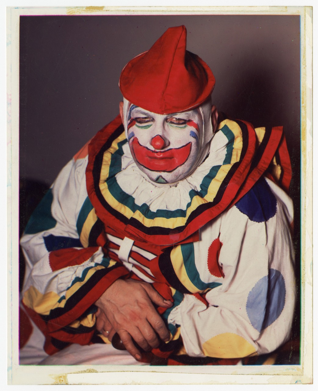 A clown wearing colorful makeup and a red costume sits with hands resting. The outfit features bold, multicolored stripes and large polka dots.