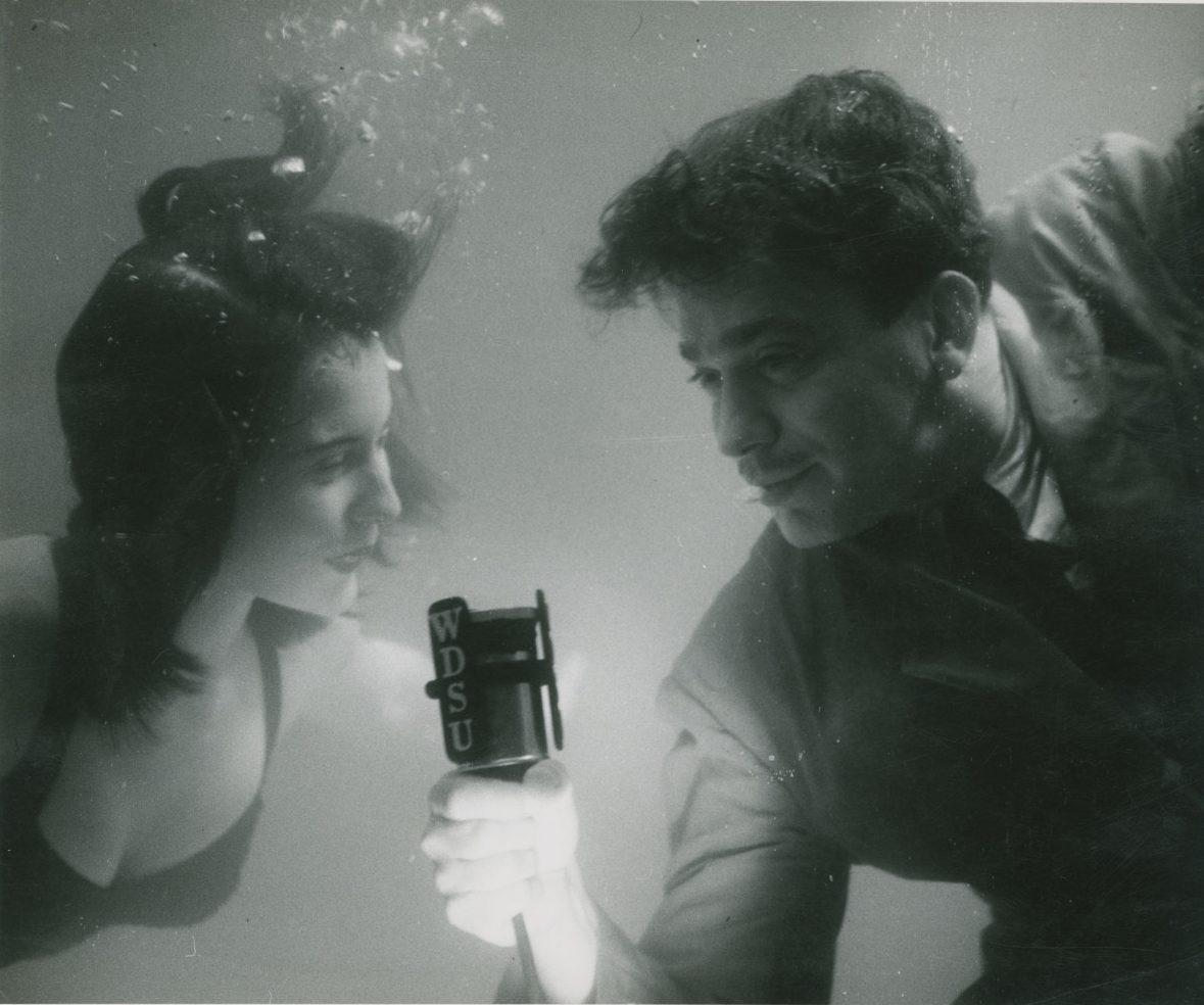 A woman and a man are underwater, looking at each other. The man is holding a microphone with WDSU printed on it. Both appear to be smiling.