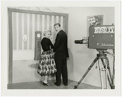 A black and white photo shows two people on a TV set, possibly in the 1960s, standing near a vintage camera. They look back at the camera, with a striped wall and framed portraits in the background.