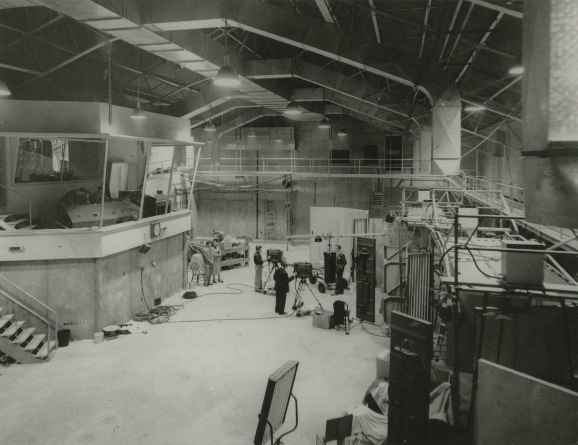 A vintage black and white photo of an industrial interior, featuring a large, high-ceilinged room with scattered equipment and a group of people working. There are scaffolds and a control booth in the background, indicating an engineering or research setting.