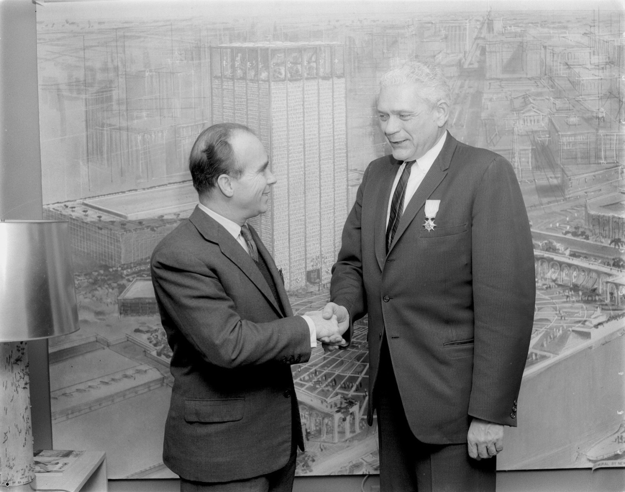 Two men in suits shake hands in front of an architectural drawing. One man is shorter and bald, the other is taller with gray hair and wears a medal on his lapel. A lamp is visible on the left side of the image.