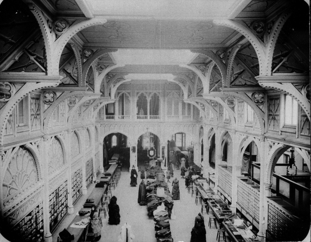 A black and white photo of a large, ornate library interior with high ceilings and decorative arches. Several people examine books and papers at long tables. Shelves line the walls, and a large, arched window is in the background.