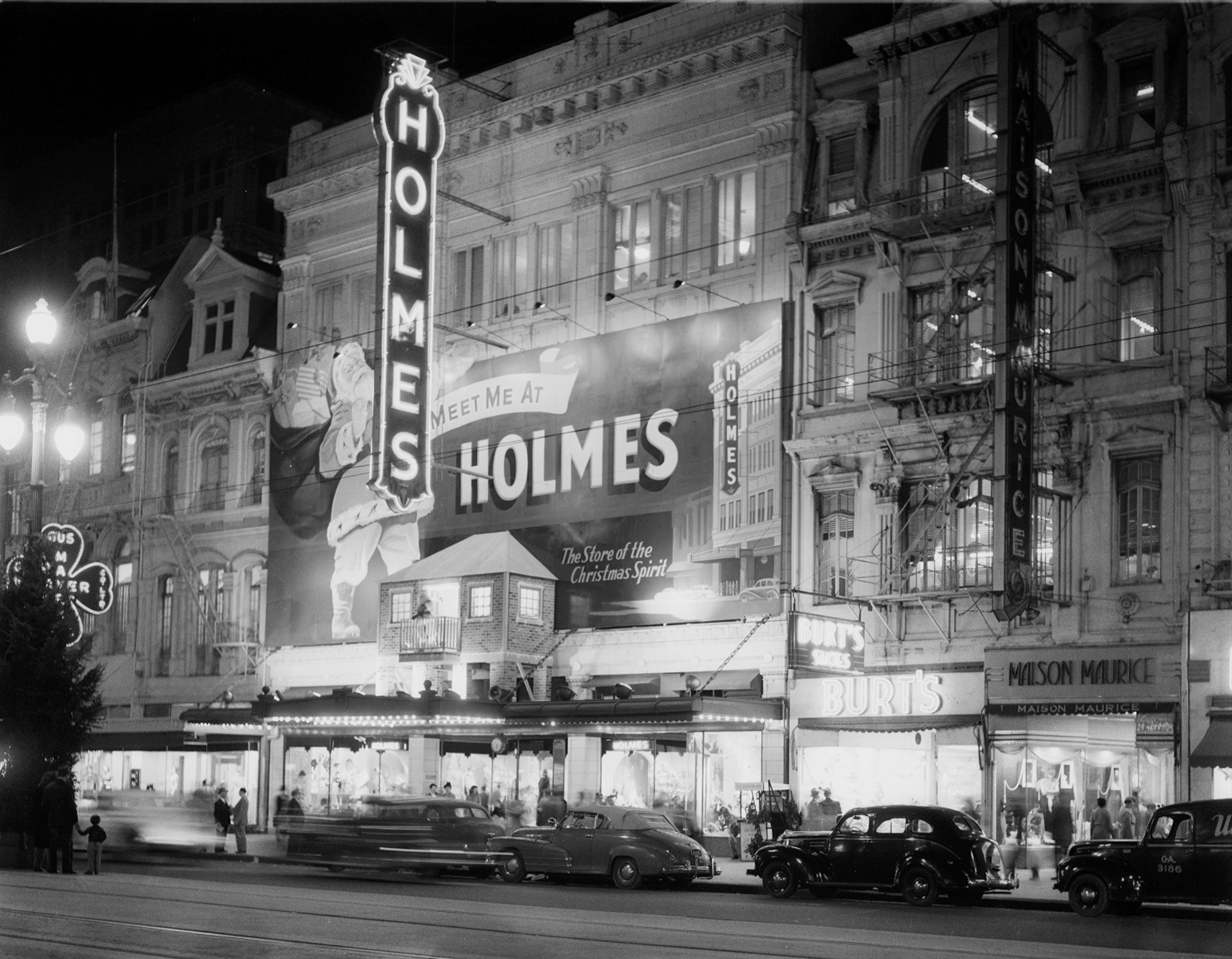 Black and white photograph of the D.H. Holmes department store in New Orleans at night. A mural on the side of the building reads: "Meet me at Holmes, the Store of the Christmas Spirit."