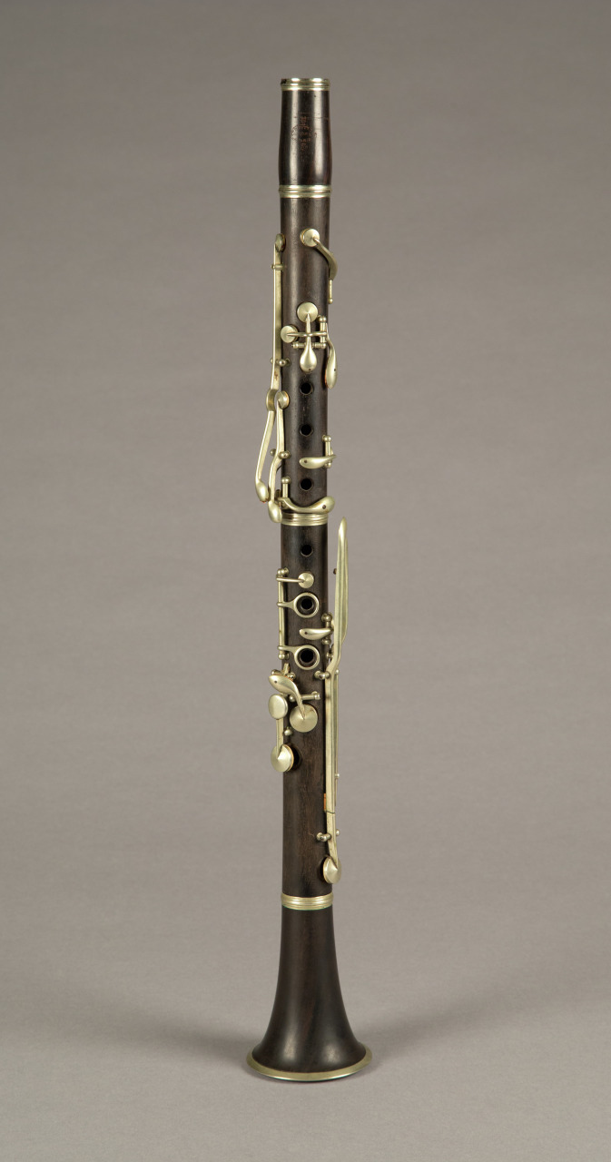 A dark wooden clarinet with silver metal keys and a smooth finish is displayed upright against a neutral gray background. The instruments bell at the bottom slightly flares out.