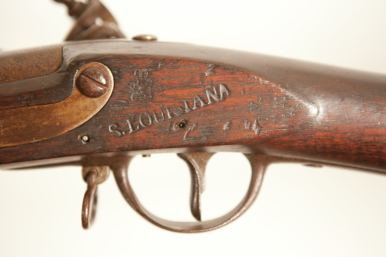 Close-up of a vintage firearm with a wooden stock and metal hardware. The word LOUISIANA is engraved on the side. The image focuses on the trigger and surrounding area, highlighting the aged, textured surface.