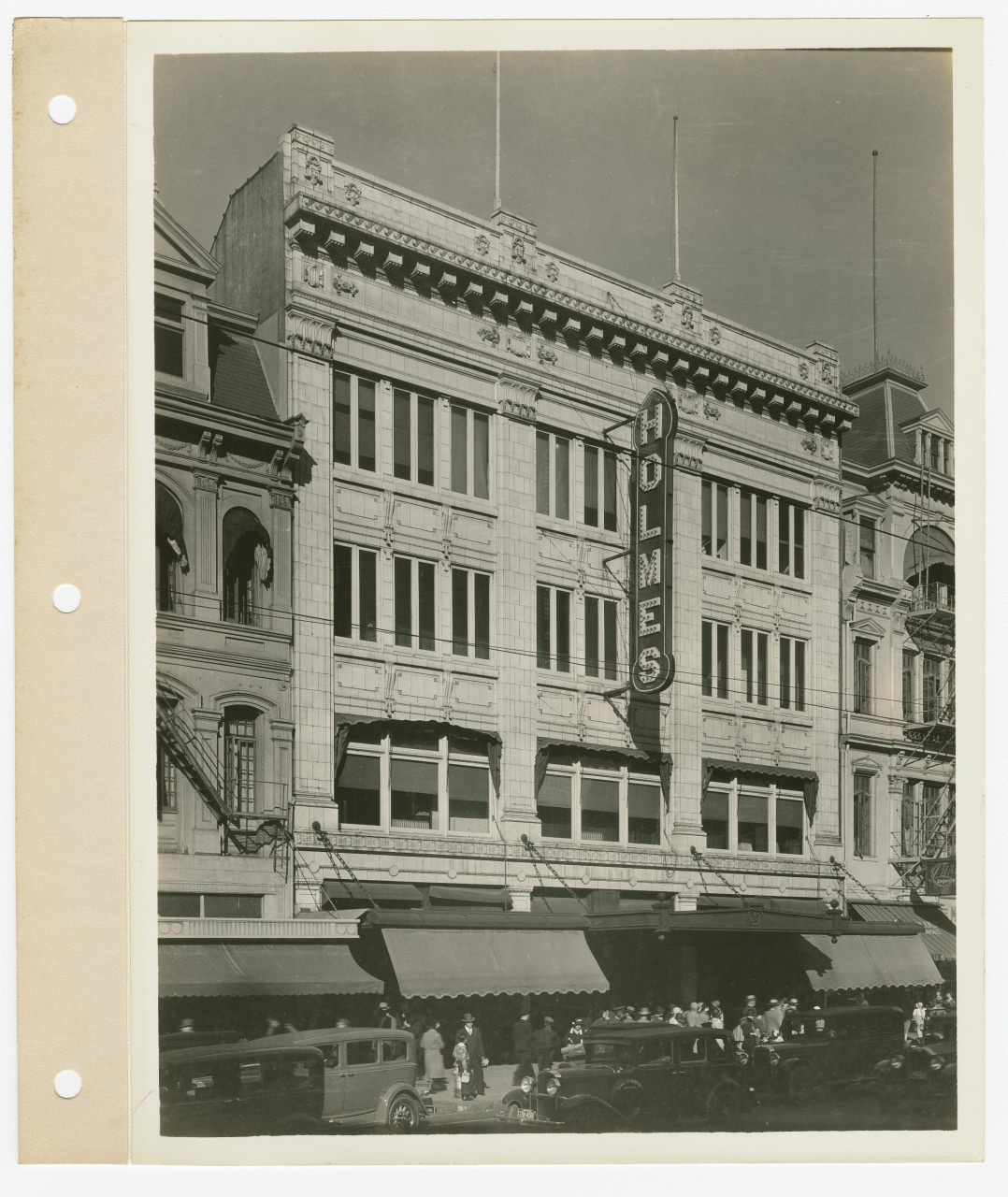 A black-and-white photo of an early 20th-century multi-story building. It features a large vertical sign reading Holmes. People and vintage cars are visible on the street in front. The architecture includes elaborate detailing.