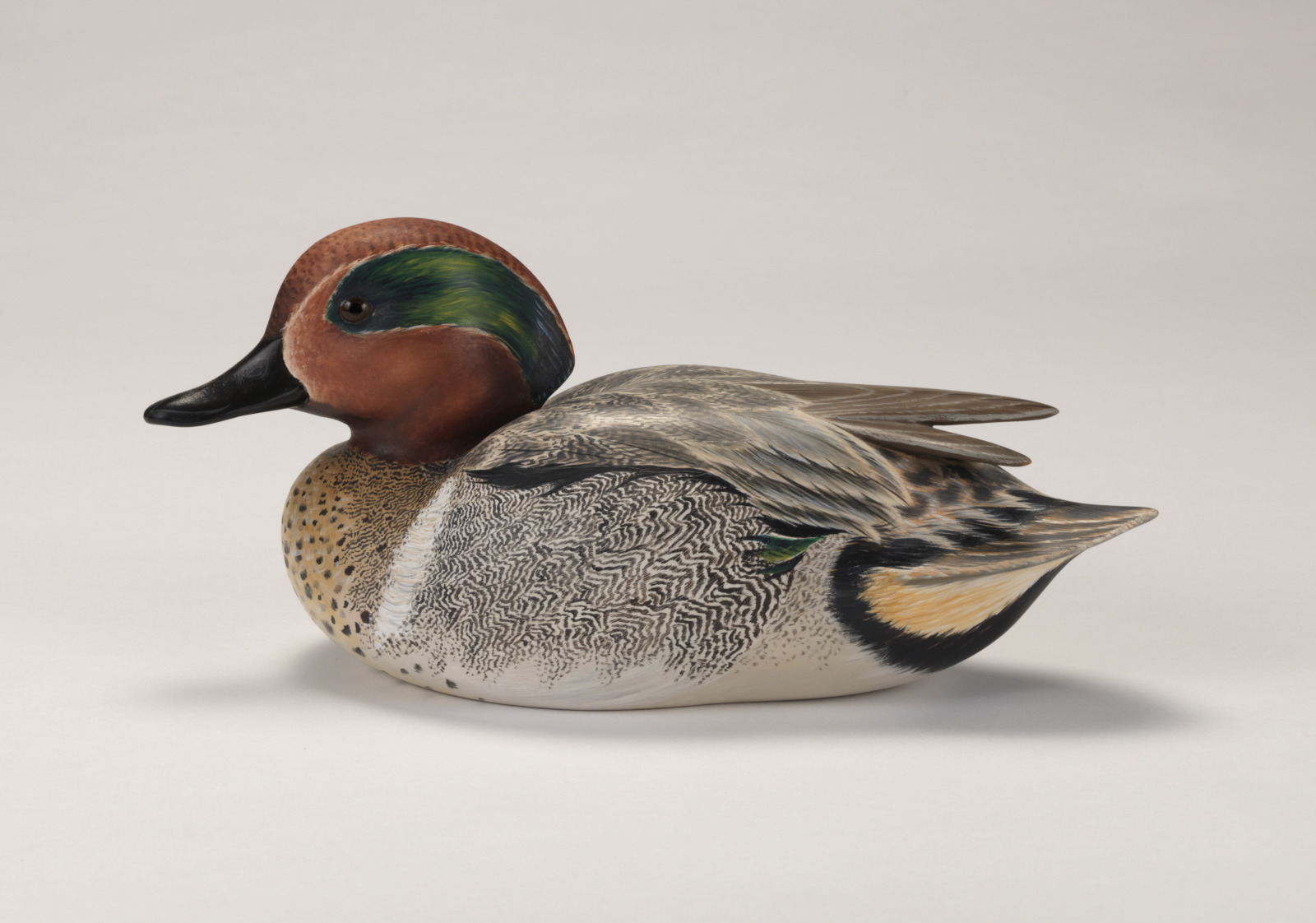 A realistic duck carving of a green wing teal depicted as if floating on water.