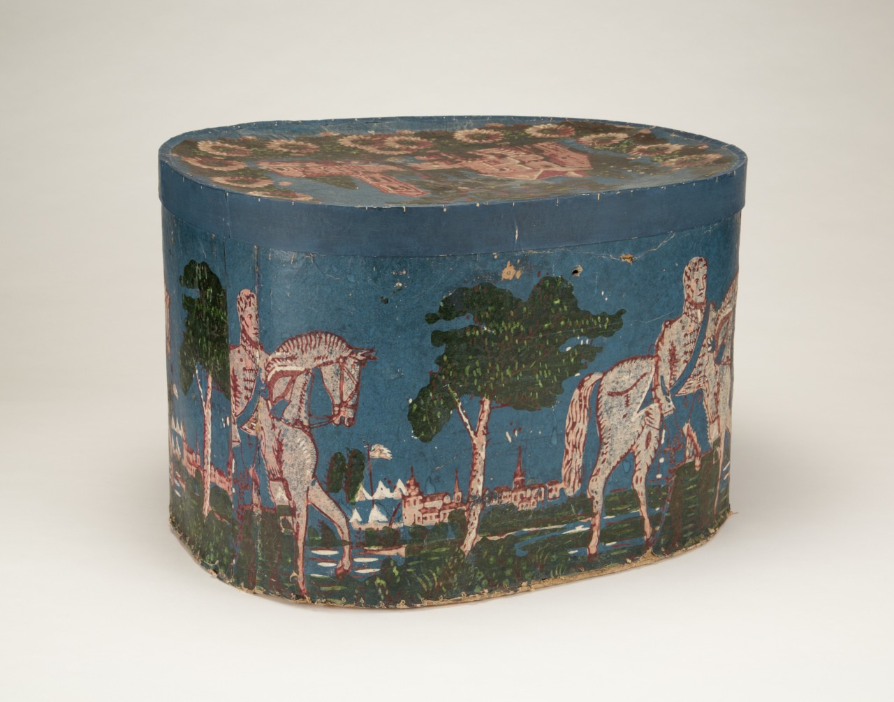 Oval hatbox with a painted scene of horses and people near trees and buildings. The background is blue with rustic details. The lid features a complementary patterned design.