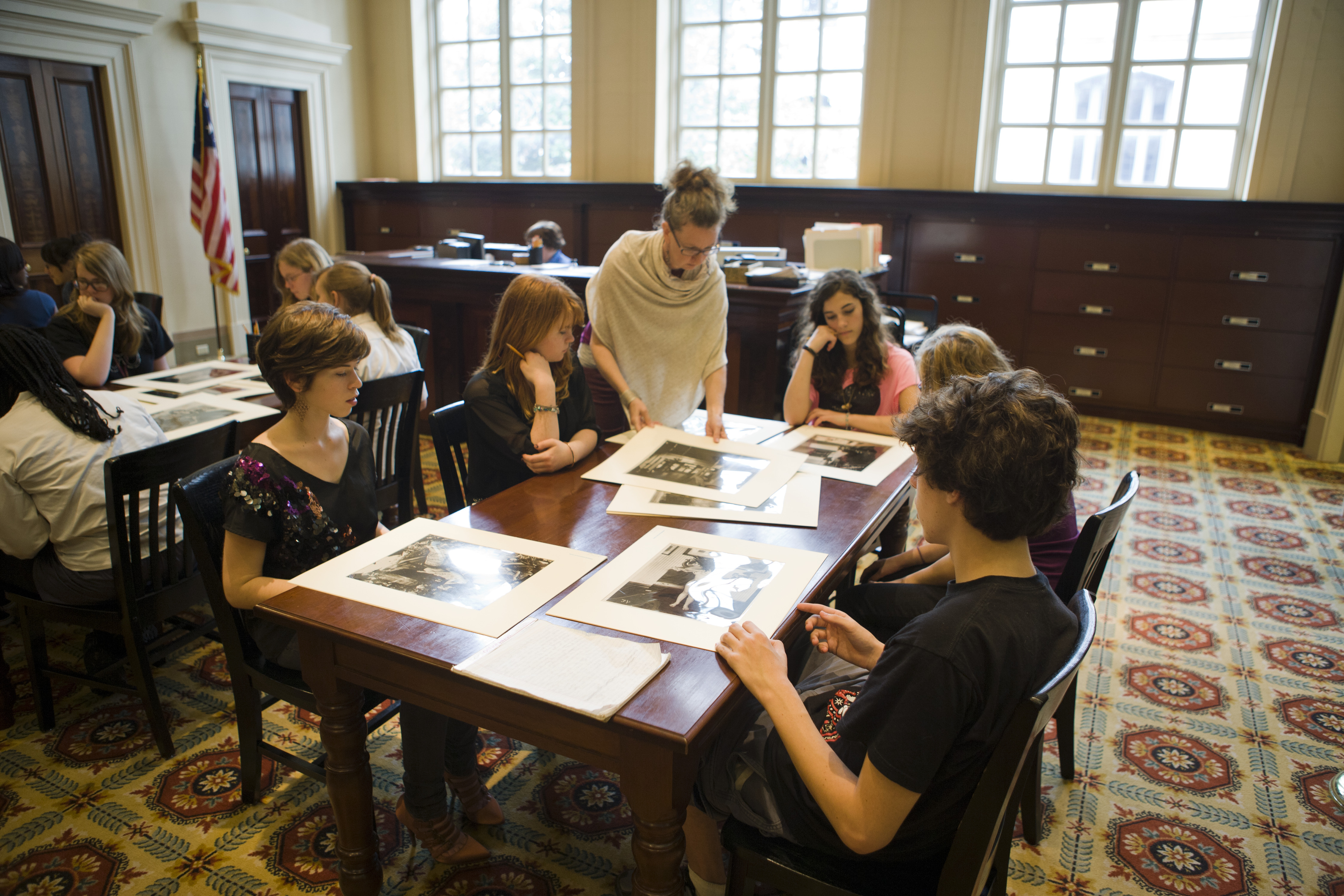 A group of people sits around a wooden table examining large black-and-white photos. The setting appears to be a library or study room with large windows and an American flag in the background. The floor has a patterned carpet.